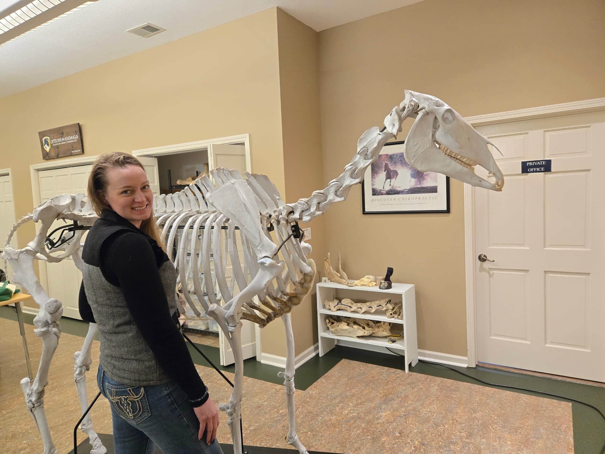 A woman stands next to a full horse skeleton in a veterinary or educational setting, smiling at the camera.